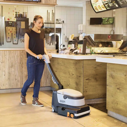 A woman in a black top and blue jeans operates a white and gray floor scrubber in a modern restaurant or cafe interior with wood-look counters and tiled walls.