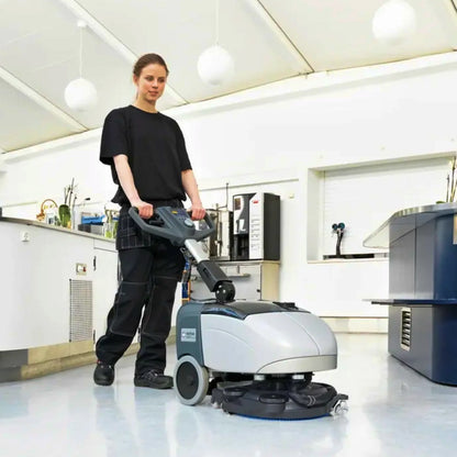 A woman in a black t-shirt and dark work pants pushes a gray and black industrial floor buffer machine in a bright, white laboratory or office space.