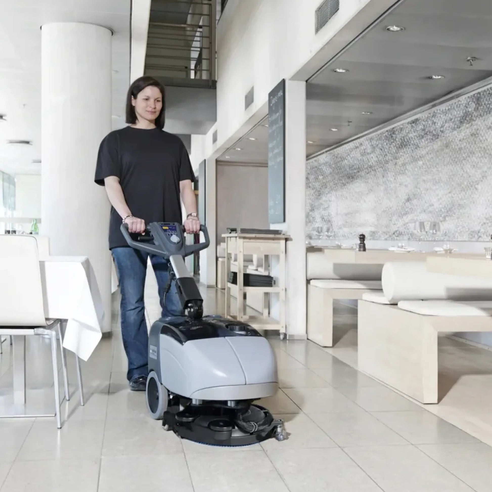 A person in a black t-shirt and jeans stands in a modern indoor lobby, holding the handle of a gray and black commercial floor buffer machine.