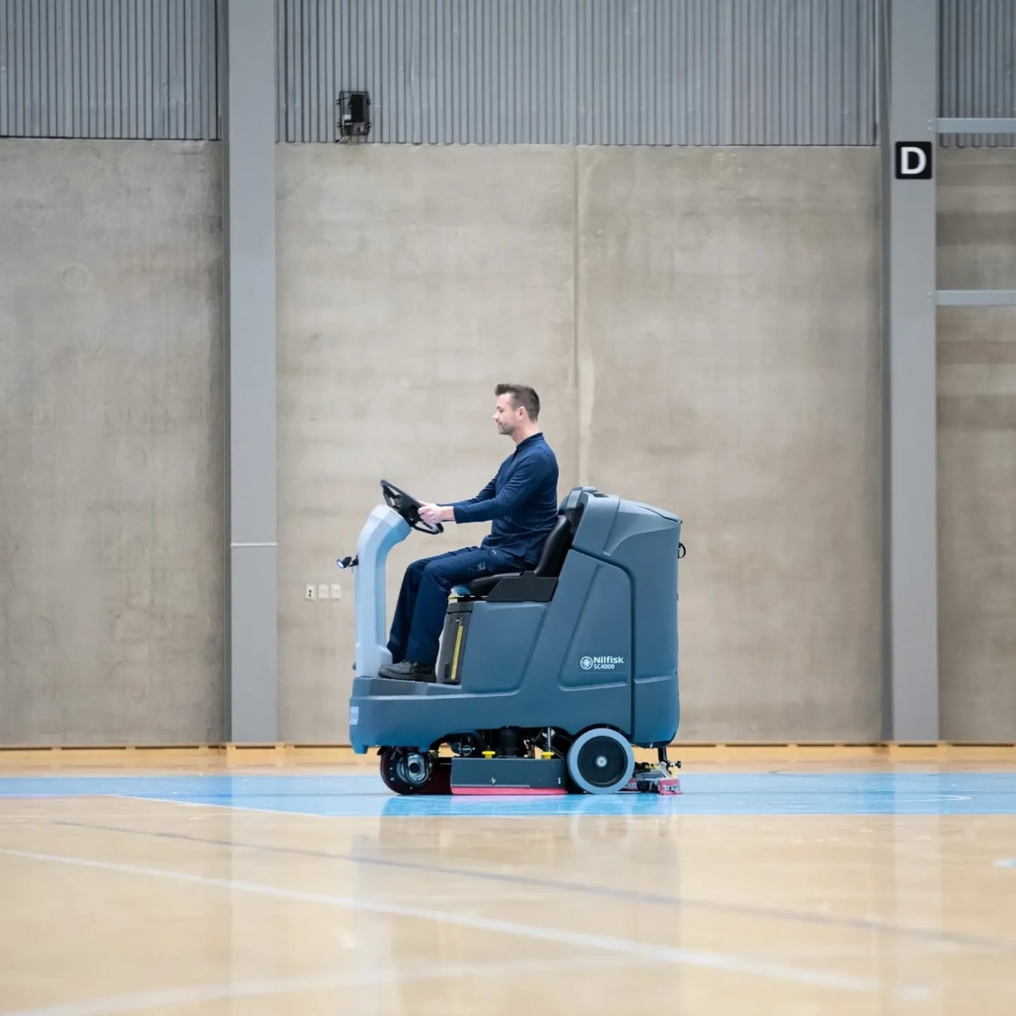 A man in a dark blue suit operates a blue and white industrial floor cleaner inside a large indoor gymnasium with a polished wood floor and concrete walls.