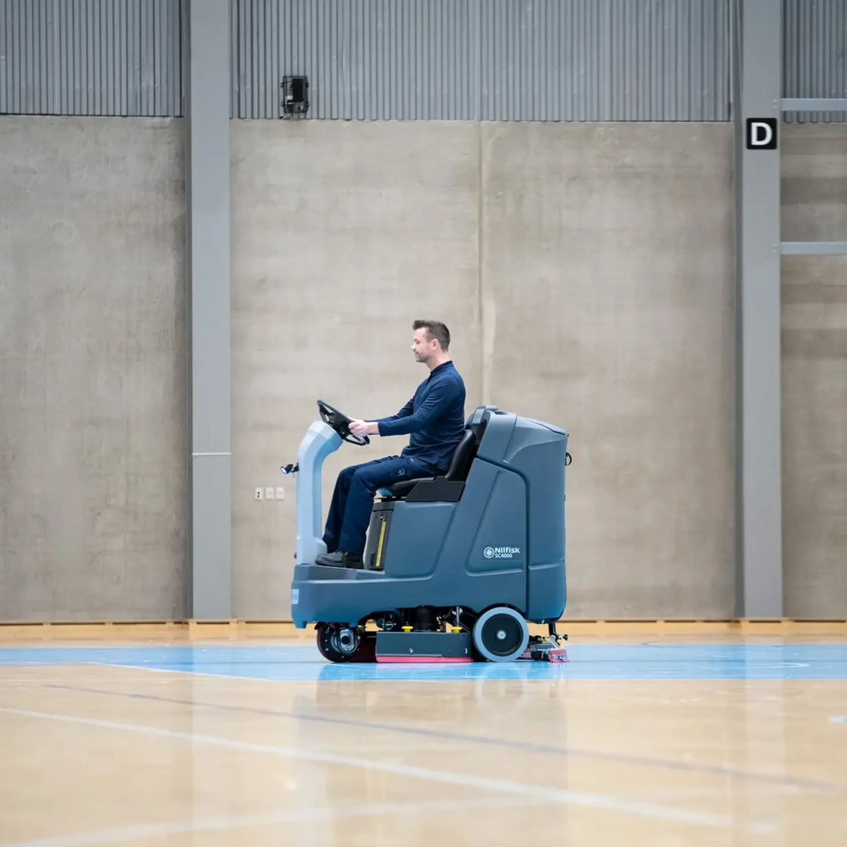 A man in a dark blue suit operates a blue and white industrial floor cleaner inside a large indoor gymnasium with a polished wood floor and concrete walls.
