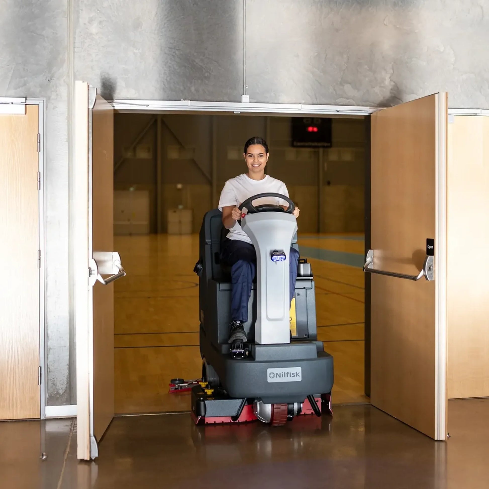 A person driving a ride-on floor scrubber inside a gymnasium, framed by open wooden doors. The machine is gray and white with red scrubbing pads visible at the bottom.