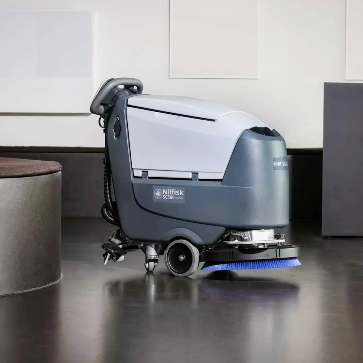 A Nilfisk IC300 floor cleaning machine with a gray and white body, shown in a real indoor environment with a dark floor and wall panels.
