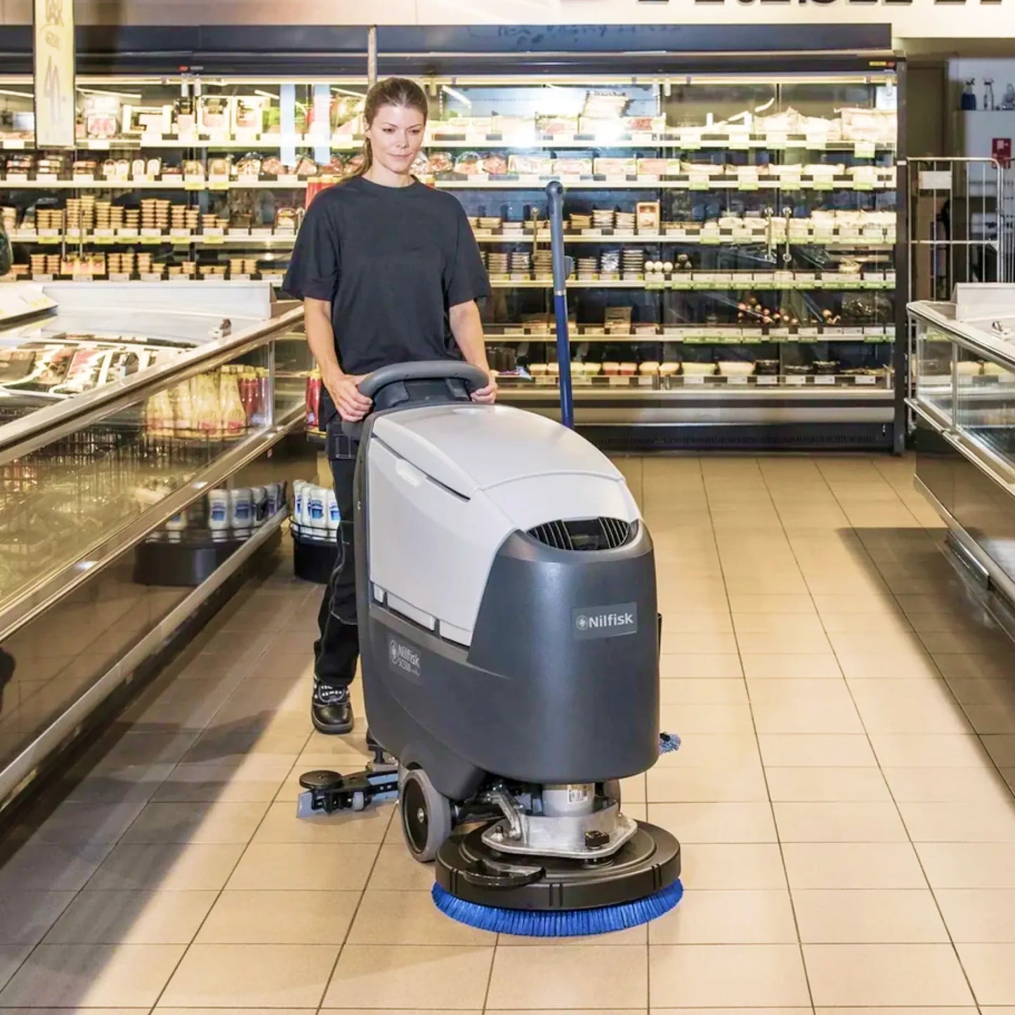 A person pushes a Kniffen commercial floor scrubber in a supermarket aisle, with tiled floors and refrigerated display cases in the background.