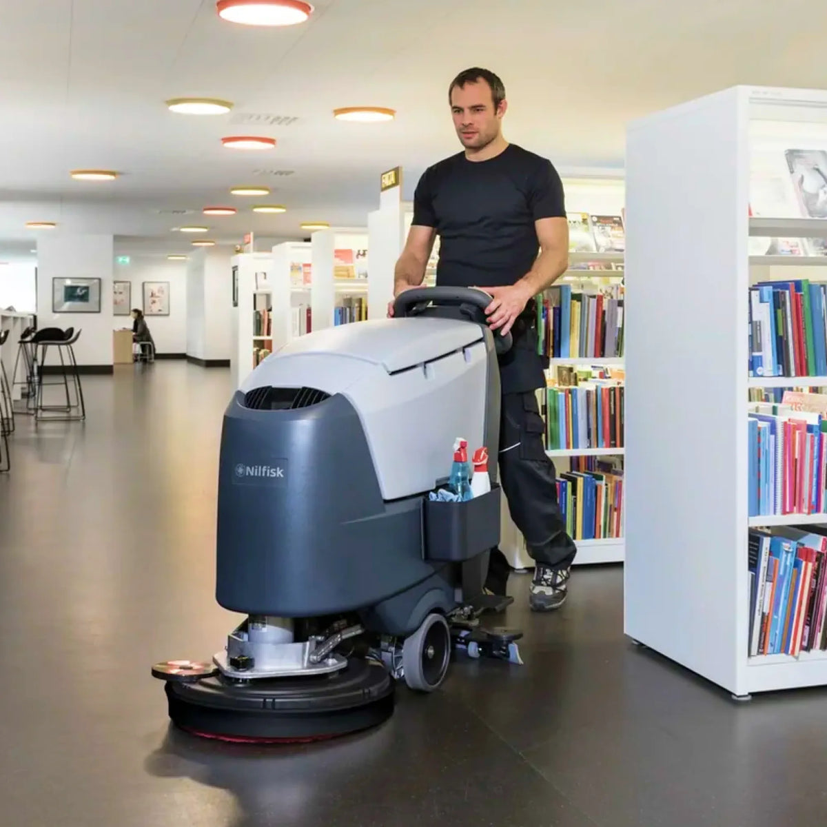 A man in a black t-shirt and dark pants pushes a Hitask floor care machine in a modern indoor library or bookstore with bookshelves visible on both sides.