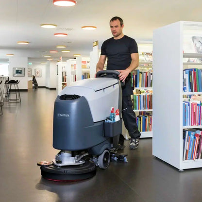 A man in a black t-shirt and dark pants pushes a Hitask floor care machine in a modern indoor library or bookstore with bookshelves visible on both sides.