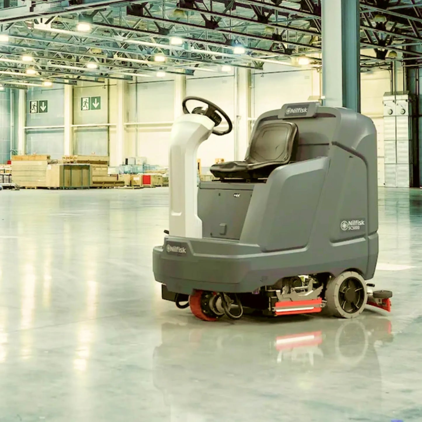 A Nittoas drive-on floor scrubber in gray and white with red accents, positioned on a polished concrete floor inside a large, brightly lit warehouse.