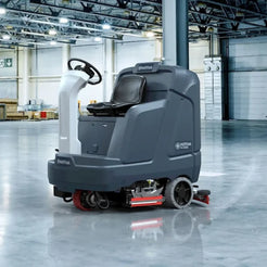 A Milftosk P120 floor care vehicle with a driver's seat, steering wheel, and brush decks, shown in a clean, spacious warehouse environment.