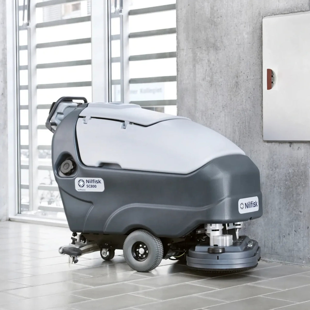 A Nilfisk drive-cleaner vacuum truck for industrial floor cleaning, shown in a modern indoor setting with tiled floors and a concrete wall.