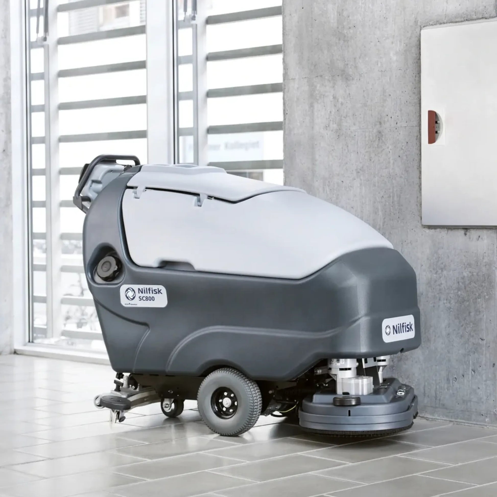 A Nilfisk drive-cleaner vacuum truck for industrial floor cleaning, shown in a modern indoor setting with tiled floors and a concrete wall.