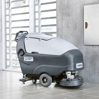 A Nilfisk drive-cleaner vacuum truck for industrial floor cleaning, shown in a modern indoor setting with tiled floors and a concrete wall.