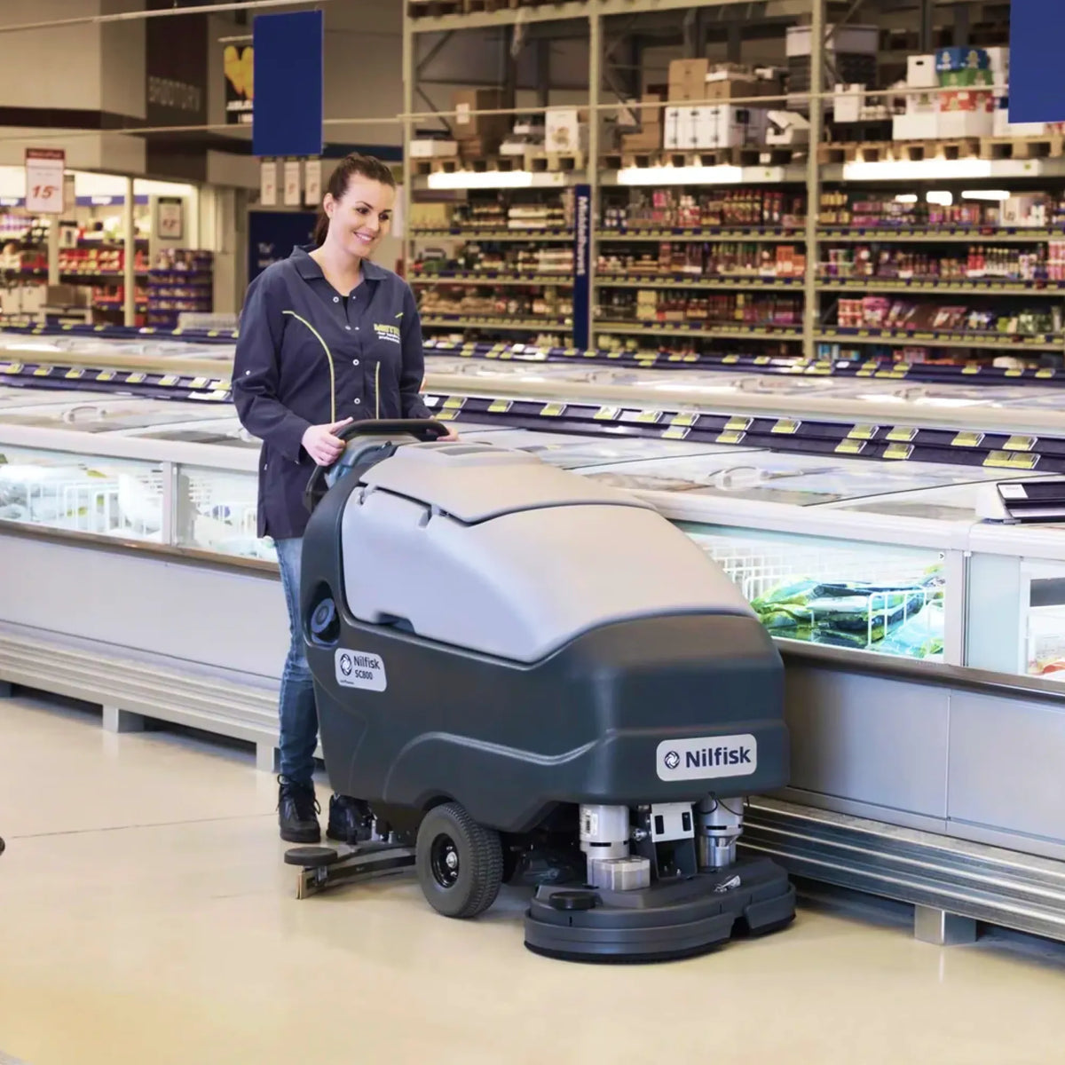 A woman wearing a dark navy uniform drives a Nilfisk commercial floor scrubber inside a supermarket aisle next to frozen food displays.