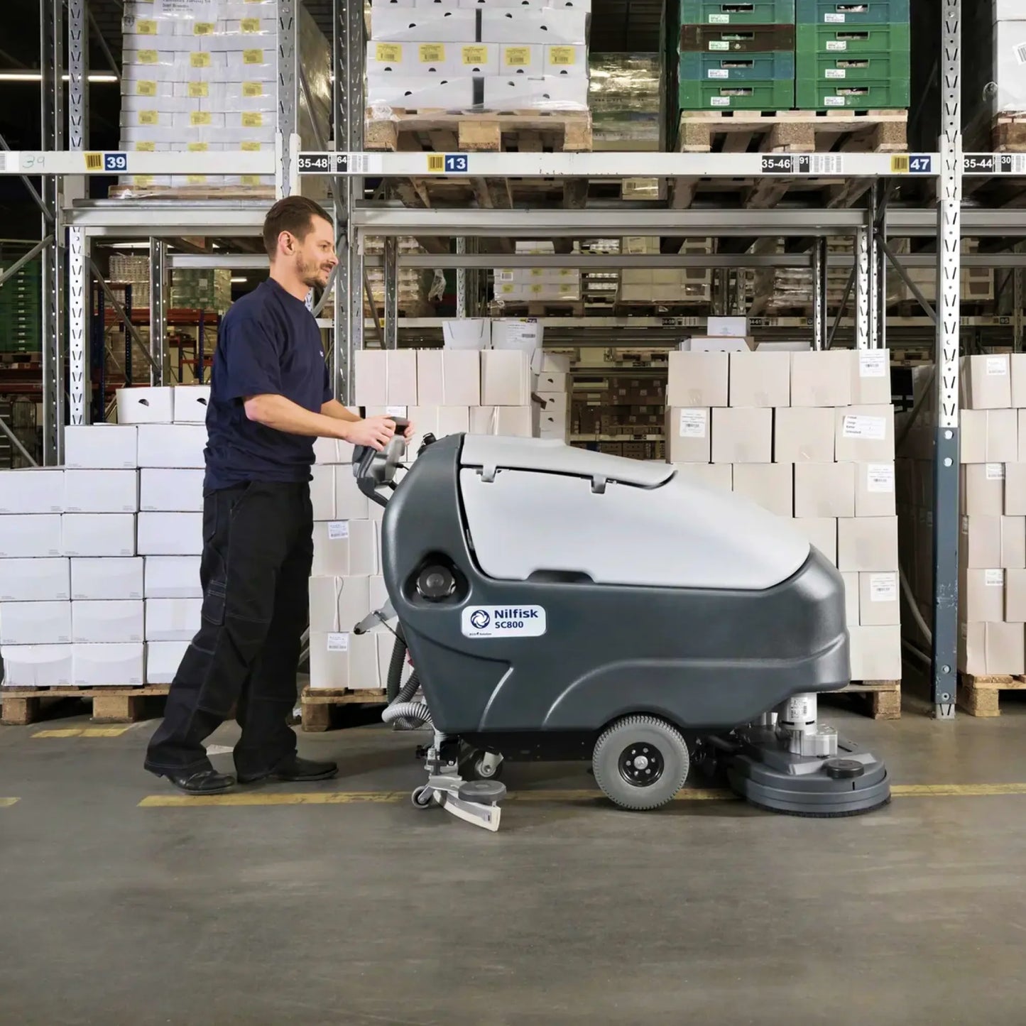 A man operating a floor-washing machine in a warehouse aisle, with shelving and stacked boxes visible in the background.
