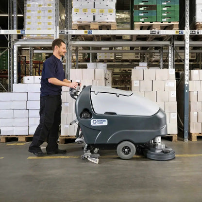 A man operating a floor-washing machine in a warehouse aisle, with shelving and stacked boxes visible in the background.