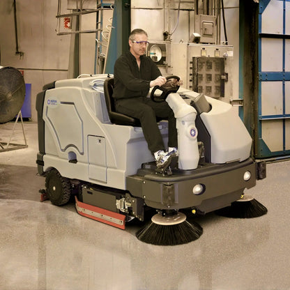 A man wearing safety glasses and a black uniform sits in the driver's seat of a white and gray industrial floor sweeper, positioned in a clean, well-lit indoor warehouse or factory environment.