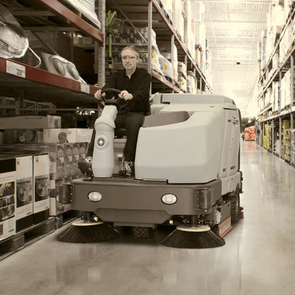 A man wearing glasses and a black shirt is driving a large gray and white industrial sweep truck aisle in a warehouse store. The warehouse shelves on both sides are stocked with various boxed and packaged goods.
