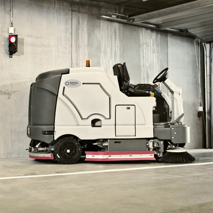 A white and gray industrial riding snowmobile with a pink stripe along the lower edge, parked inside a concrete warehouse or garage area. The side of the machine shows the 'NexGen' branding.
