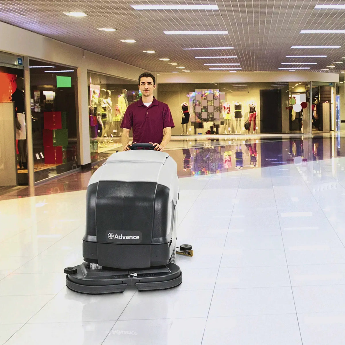 A man in a maroon polo shirt operates an Advance brand commercial floor scrubber in a brightly lit, modern shopping mall corridor with white tiled floors.