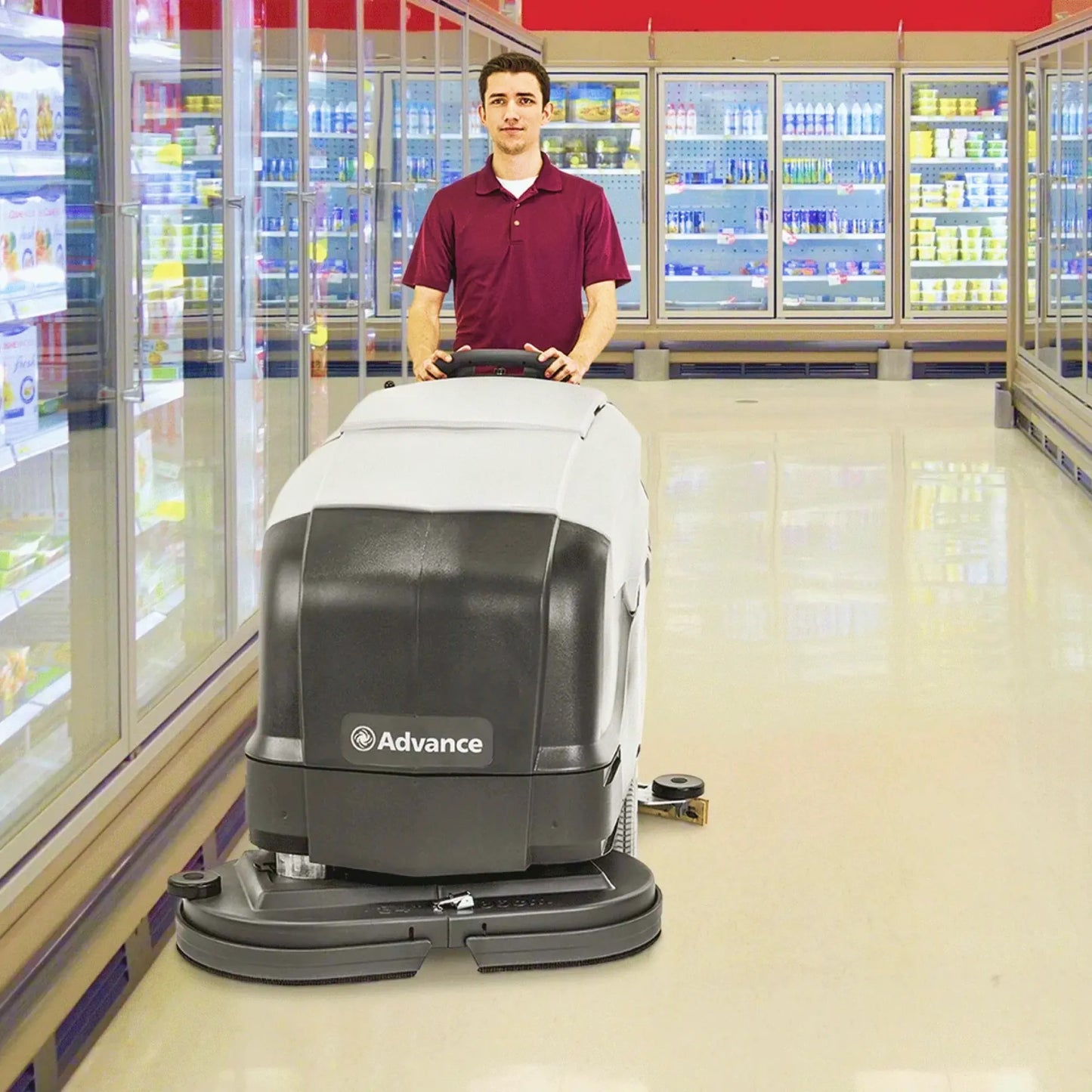 A man in a maroon polo shirt operates an Advance brand commercial floor scrubber in a brightly lit supermarket aisle with glass-door beverage refrigerators in the background.